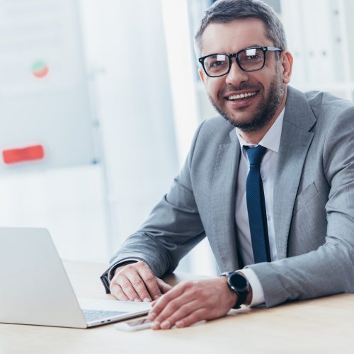 cheerful businessman in eyeglasses using laptop and smiling at camera in office cheerful businessman in eyeglasses using laptop and smiling at camera in office